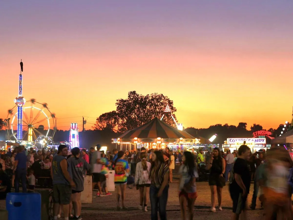 The Stoddard County Fair midway at sunset, with a lit Ferris wheel, carousel and cotton candy stand, and a crowd walking the fairgrounds.