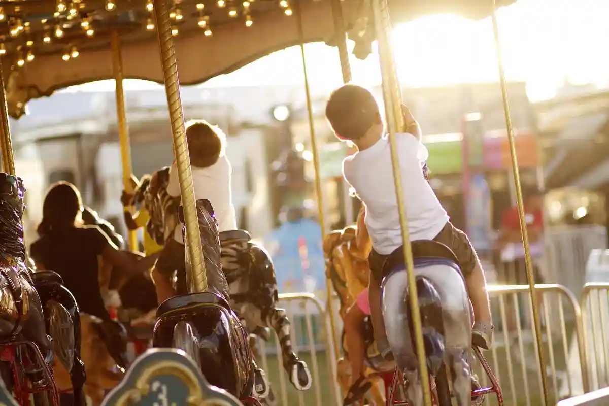 Two children riding a lit carousel at a county fair, reaching for the horses' reins as the sun sets behind them.